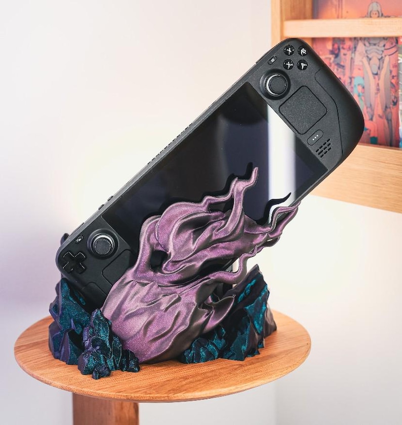 May include: A black handheld gaming device with a purple and blue 3D printed stand shaped like flames. The stand is on a wooden base.