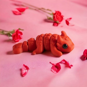 May include: A handcrafted, orange-red polymer clay creature with a segmented body and large, black eyes. The sculpture is posed on a pink satin surface, with red rose petals and roses in the background.