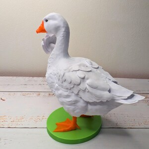 May include: A white goose ornament with an orange beak and feet, standing on a green circular base. The goose has detailed feather textures and is posed in a side profile.