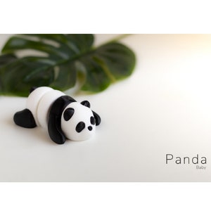 May include: A small, black and white panda figurine lying on its belly. The panda is made of a smooth, white material with black markings around its eyes and ears. The panda is on a white surface with a green leaf in the background.