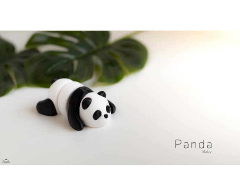 May include: A small, black and white panda figurine lying on its belly. The panda is made of a smooth, white material with black markings around its eyes and ears. The panda is on a white surface with a green leaf in the background.