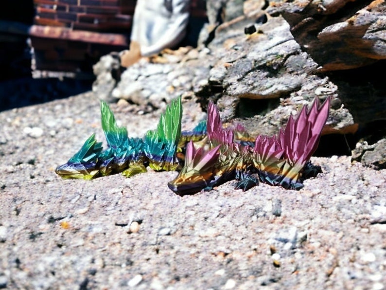 May include: Two iridescent crystal lizards, one green and one purple, are lying on a sandy surface. The lizards are made of faceted crystals and have a metallic sheen.