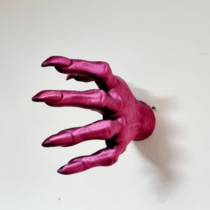 Puede incluir: Un gancho de pared con forma de mano de color rosa oscuro con dedos largos y puntiagudos.