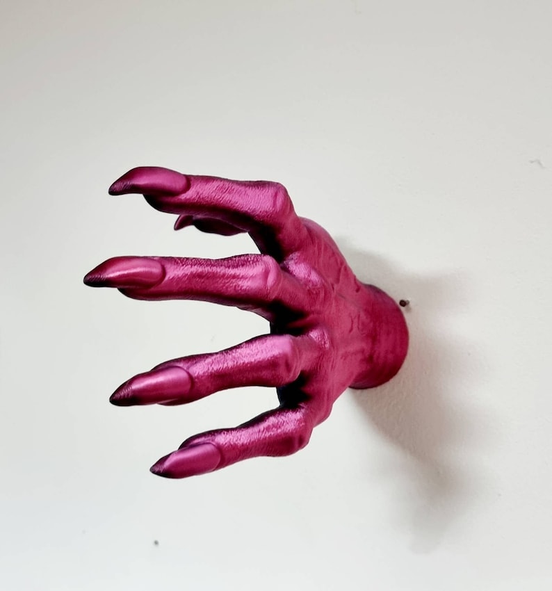 Puede incluir: Un gancho de pared con forma de mano de color rosa oscuro con dedos largos y puntiagudos.