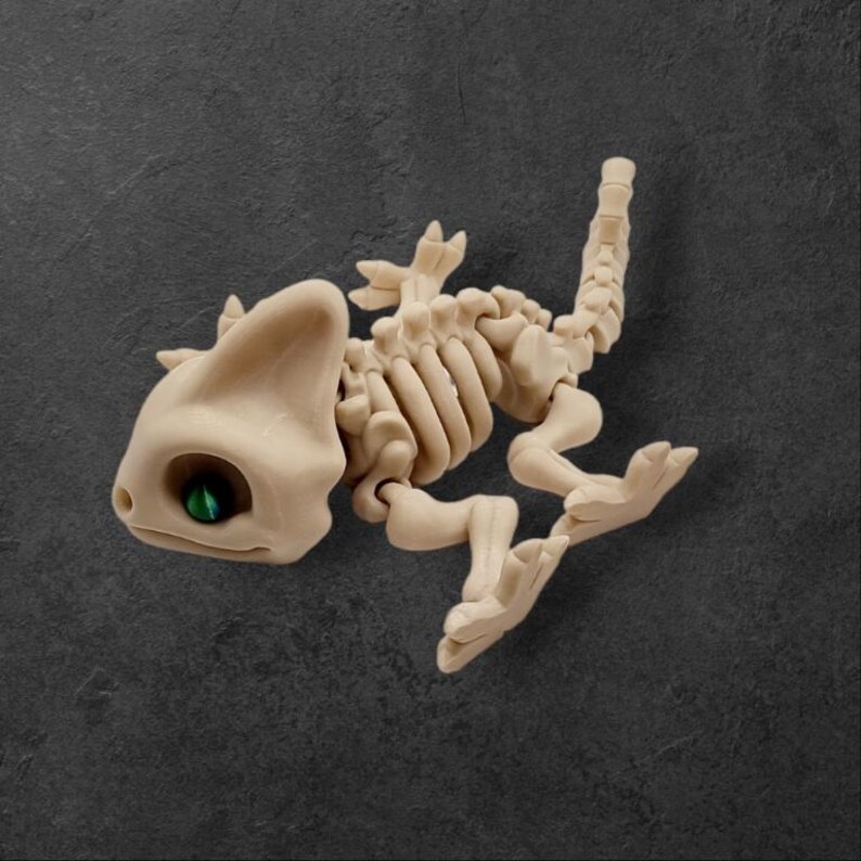 May include: A beige plastic skeleton of a chameleon with a green eye. The chameleon is posed in a running position with its tail curled up.