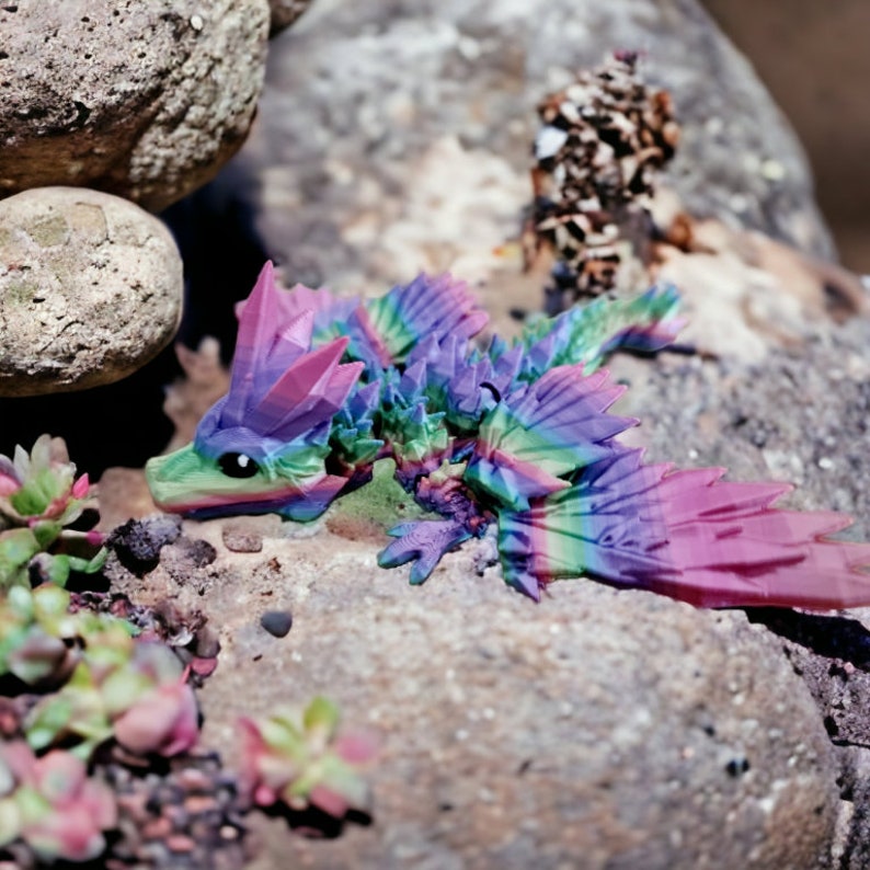 K&ouml;nnte beinhalten: Eine farbenfrohe, vielseitige, irisierende Drachenfigur mit rosa, lila, blauen und gr&uuml;nen Akzenten. Der Drache ist mit ausgebreiteten Fl&uuml;geln und seitlich gedrehtem Kopf dargestellt. Die Figur ist aus einem glatten, harten Material gefertigt und hat eine gl&auml;nzende Oberfl&auml;che.