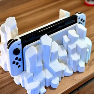 May include: A white Nintendo Switch console is displayed in a white, 3D printed stand that resembles a rocky mountain range.