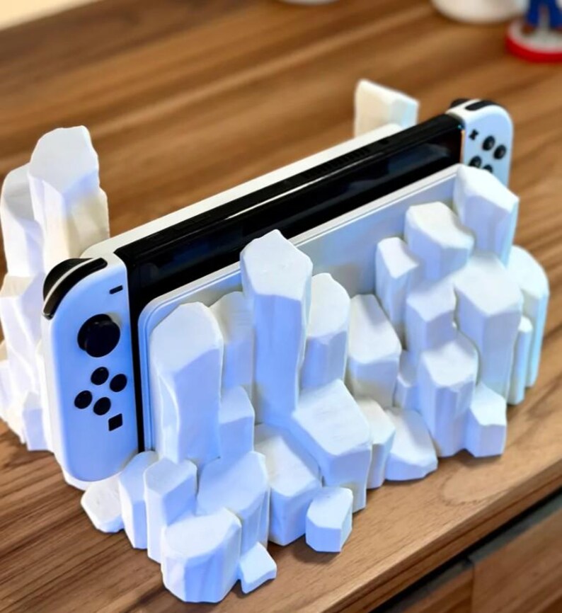 May include: A white Nintendo Switch console is displayed in a white, 3D printed stand that resembles a rocky mountain range.