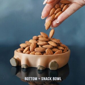 Op de afbeelding: Een bruine snackkom gevuld met amandelen, met meer amandelen die van bovenaf worden ingegoten. De kom heeft een uniek ontwerp met vier kleine voetjes en de tekst "BOTTOM = SNACK BOWL" onderaan.