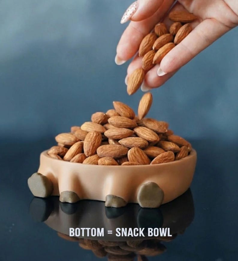 Op de afbeelding: Een bruine snackkom gevuld met amandelen, met meer amandelen die van bovenaf worden ingegoten. De kom heeft een uniek ontwerp met vier kleine voetjes en de tekst "BOTTOM = SNACK BOWL" onderaan.