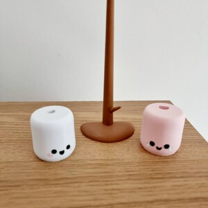 Op de afbeelding: Twee kleine, ronde, marshmallow-vormige figuurtjes met glimlachende gezichten. De witte marshmallow is links en de roze marshmallow is rechts. Een bruine houten stok staat in het midden.