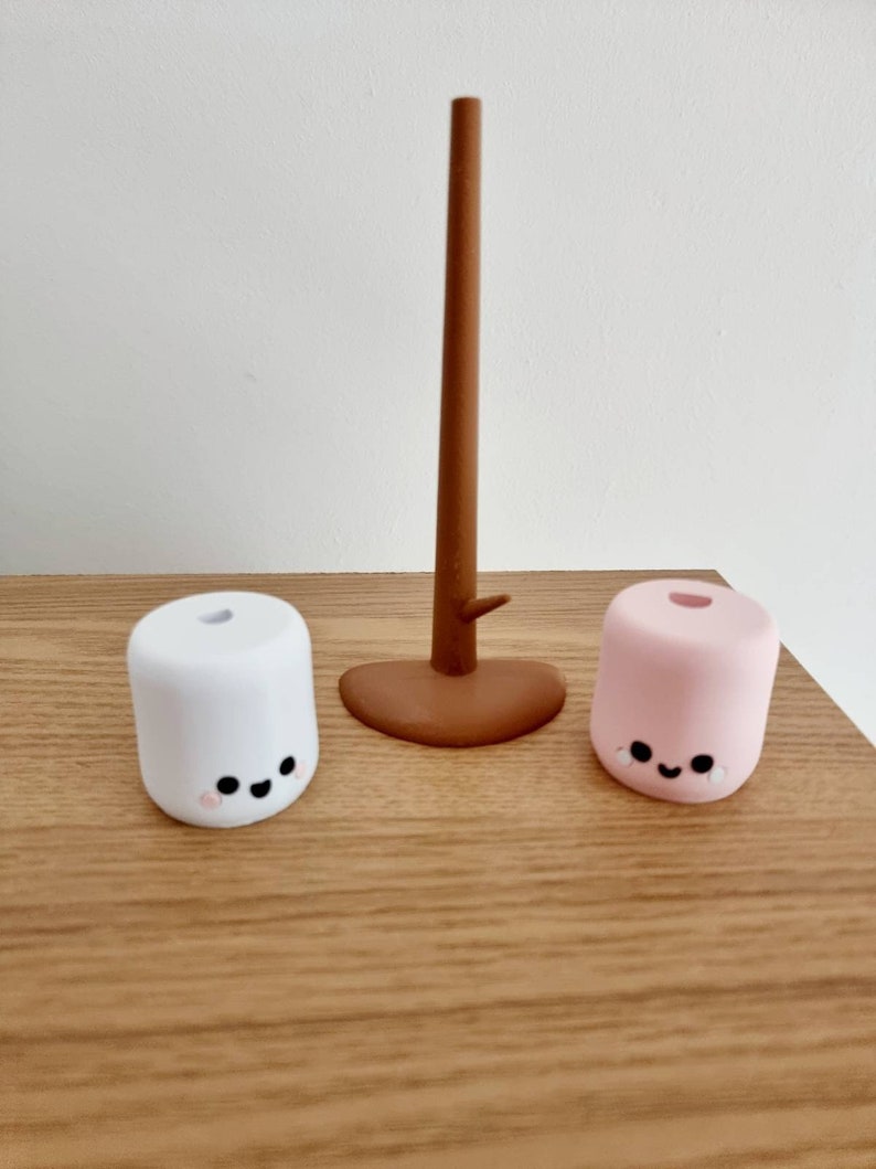 Op de afbeelding: Twee kleine, ronde, marshmallow-vormige figuurtjes met glimlachende gezichten. De witte marshmallow is links en de roze marshmallow is rechts. Een bruine houten stok staat in het midden.
