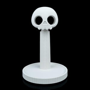May include: A white, decorative paper towel holder with a skull-shaped top. The skull has large eye sockets and a detailed lower jaw. The holder has a cylindrical stem and a round base with a grooved edge. The design is minimalist and modern.