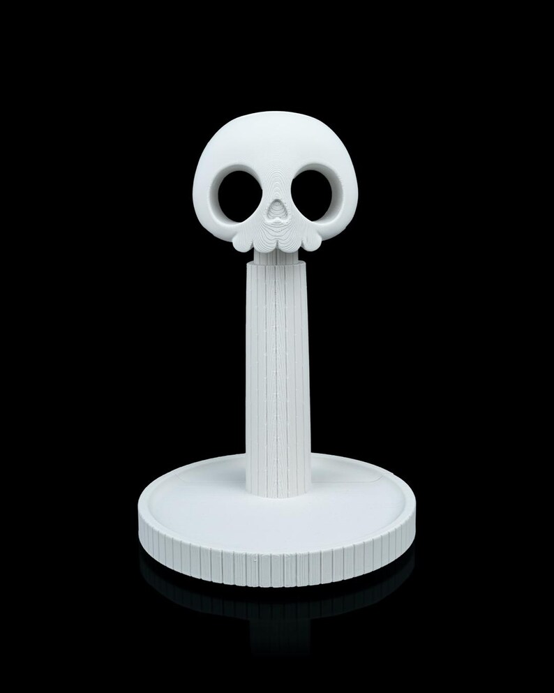 May include: A white, decorative paper towel holder with a skull-shaped top. The skull has large eye sockets and a detailed lower jaw. The holder has a cylindrical stem and a round base with a grooved edge. The design is minimalist and modern.