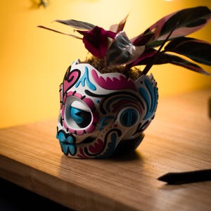 May include: A decorative skull-shaped planter, painted white with pink and blue accents, holds a houseplant with pink and green leaves. The planter sits on a wooden surface against a yellow background.