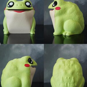 May include: A whimsical, light green frog-shaped container with a wide, open mouth and large, expressive eyes. The frog has a cream-colored belly and red accents on its cheeks and mouth. The container is designed to hold small items.
