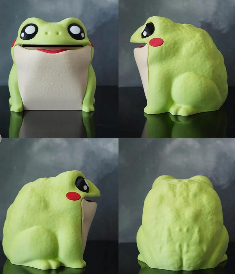 May include: A whimsical, light green frog-shaped container with a wide, open mouth and large, expressive eyes. The frog has a cream-colored belly and red accents on its cheeks and mouth. The container is designed to hold small items.