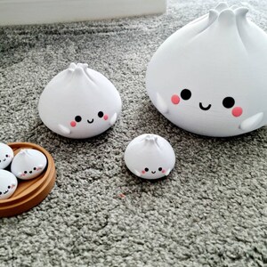 May include: A set of five white ceramic dumplings with happy faces. The dumplings are in different sizes and are arranged in a wooden steamer basket. The largest dumpling is standing on its own, while the smaller dumplings are inside the basket.