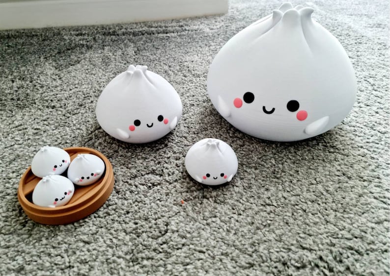 May include: A set of five white ceramic dumplings with happy faces. The dumplings are in different sizes and are arranged in a wooden steamer basket. The largest dumpling is standing on its own, while the smaller dumplings are inside the basket.