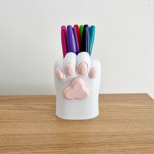 May include: White ceramic paw-shaped pencil holder with pink paw pads, holding a variety of colored pens.