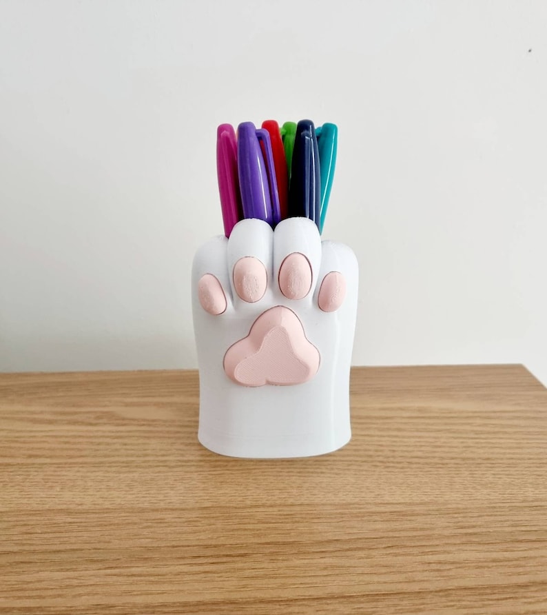 May include: White ceramic paw-shaped pencil holder with pink paw pads, holding a variety of colored pens.