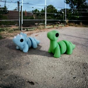 Puede incluir: Dos peque&ntilde;os juguetes de dinosaurio segmentados. Uno es azul claro y el otro es verde. Ambos est&aacute;n colocados sobre una superficie gris y arenosa.