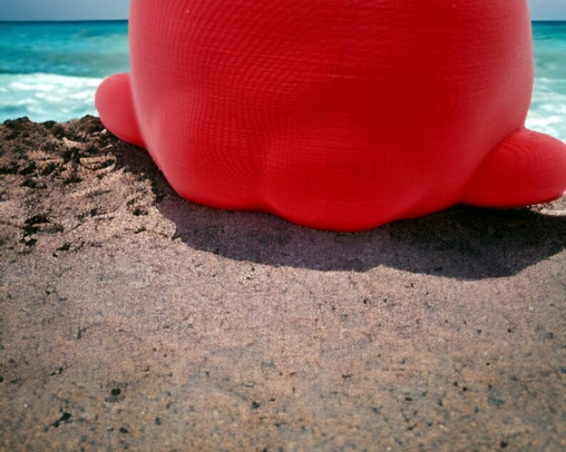 May include: A bright red inflatable toy shaped like a large, round, and smooth object. The toy is sitting on a sandy beach.