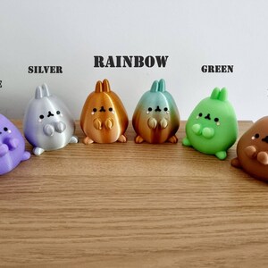 K&ouml;nnte beinhalten: Sechs kleine, pl&uuml;schige Hasenfiguren in verschiedenen Farben, darunter lila, silber, regenbogen, gr&uuml;n und braun. Die Figuren sind in einer Reihe auf einer Holzoberfl&auml;che angeordnet. Die W&ouml;rter "PURPLE", "SILVER", "RAINBOW", "GREEN" und "BROWN" stehen &uuml;ber jeder Figur.