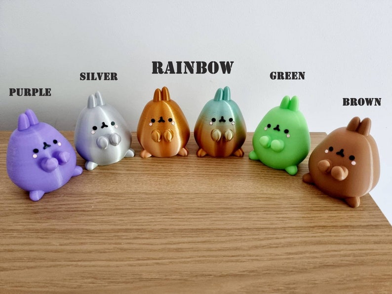 K&ouml;nnte beinhalten: Sechs kleine, pl&uuml;schige Hasenfiguren in verschiedenen Farben, darunter lila, silber, regenbogen, gr&uuml;n und braun. Die Figuren sind in einer Reihe auf einer Holzoberfl&auml;che angeordnet. Die W&ouml;rter "PURPLE", "SILVER", "RAINBOW", "GREEN" und "BROWN" stehen &uuml;ber jeder Figur.