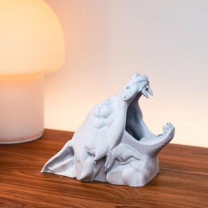 Puede incluir: Una escultura blanca impresa en 3D de una criatura con la boca abierta, revelando dientes afilados. La escultura est&aacute; sentada sobre una superficie de madera.