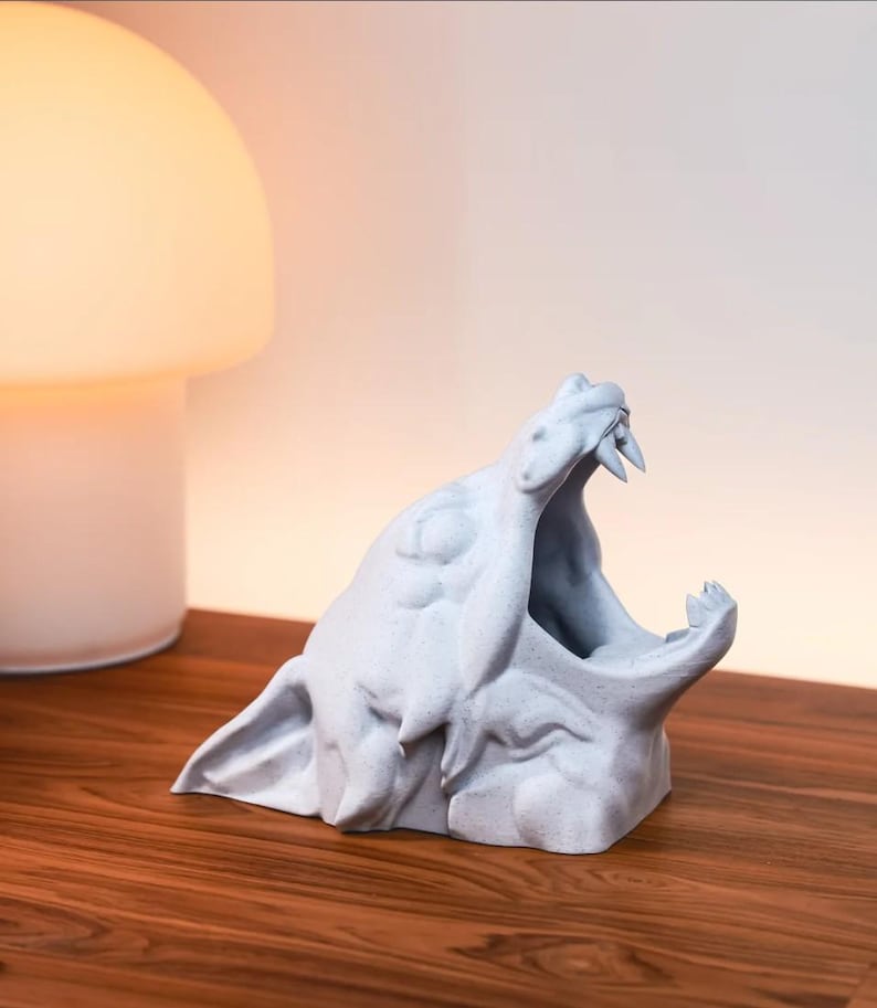 Puede incluir: Una escultura blanca impresa en 3D de una criatura con la boca abierta, revelando dientes afilados. La escultura est&aacute; sentada sobre una superficie de madera.