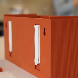 May include: An orange rectangular box with vertical ribbed texture. The box has two white strips attached to the side and a small hole. The box is sitting on a white surface.