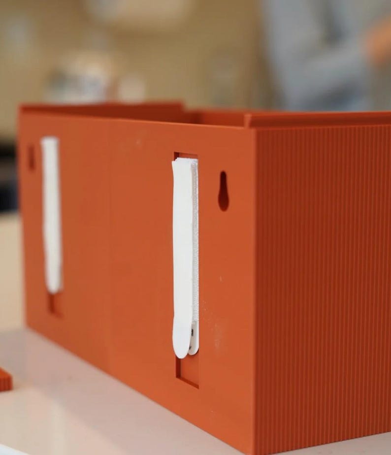 May include: An orange rectangular box with vertical ribbed texture. The box has two white strips attached to the side and a small hole. The box is sitting on a white surface.