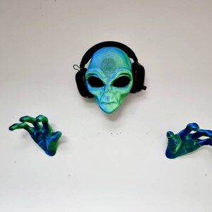 May include: A green and blue alien mask with black headphones on a white background. The alien has large eyes and a serious expression. Two green and blue alien hands are on either side of the mask.