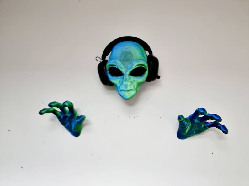 May include: A green and blue alien mask with black headphones on a white background. The alien has large eyes and a serious expression. Two green and blue alien hands are on either side of the mask.