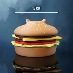 Op de afbeelding: Een hamburger-vormige container, ongeveer 13 cm in diameter, met een sesamzaadje broodje bovenop. De burger heeft lagen bruin, geel, rood en groen, die een klassieke burger nabootsen. De container wordt weerspiegeld in een glanzend oppervlak.