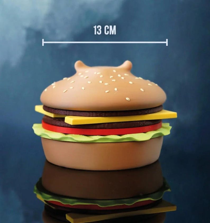Op de afbeelding: Een hamburger-vormige container, ongeveer 13 cm in diameter, met een sesamzaadje broodje bovenop. De burger heeft lagen bruin, geel, rood en groen, die een klassieke burger nabootsen. De container wordt weerspiegeld in een glanzend oppervlak.
