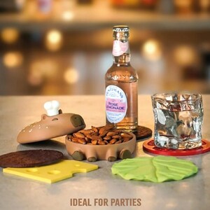 Op de afbeelding: Een grillige feestopstelling met een capibara-vormige snackkom met amandelen, een fles rozenlimonade en een getextureerd glas. Inclusief burger-vormige onderzetters: een broodje, kaas en sla.