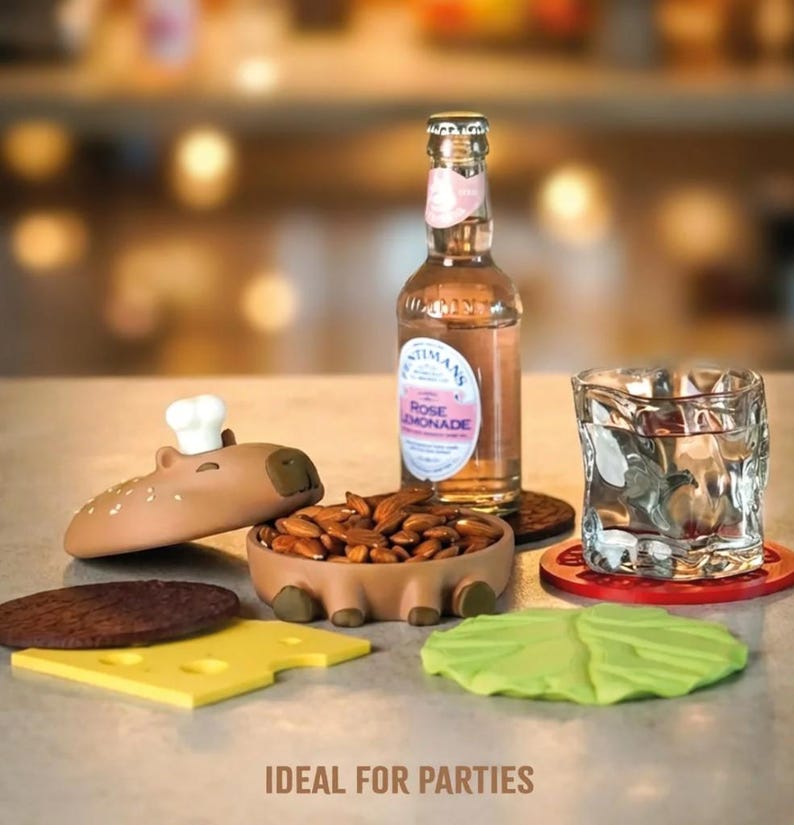 Op de afbeelding: Een grillige feestopstelling met een capibara-vormige snackkom met amandelen, een fles rozenlimonade en een getextureerd glas. Inclusief burger-vormige onderzetters: een broodje, kaas en sla.