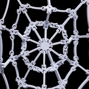 May include: A white plastic spider web model with a circular center and radiating spokes. The web is made of interlocking pieces that create a delicate and intricate design.