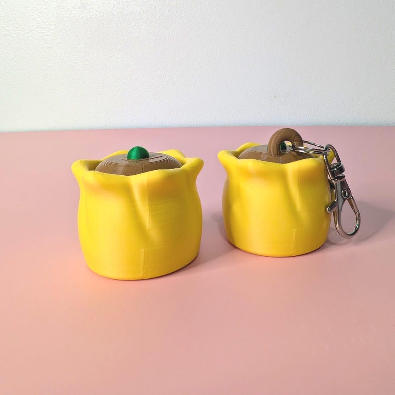 May include: Two yellow pouches with brown tops and green accents. One has a silver keychain attached. The pouches are sitting on a pink surface. The pouches have a rounded shape.