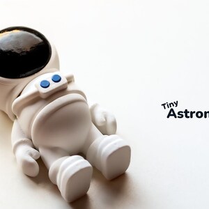 May include: A white astronaut figurine with a black helmet and blue details. The figurine is lying on its back with its arms and legs outstretched. The text "Tiny Astronaut" is written in black letters on a white background.
