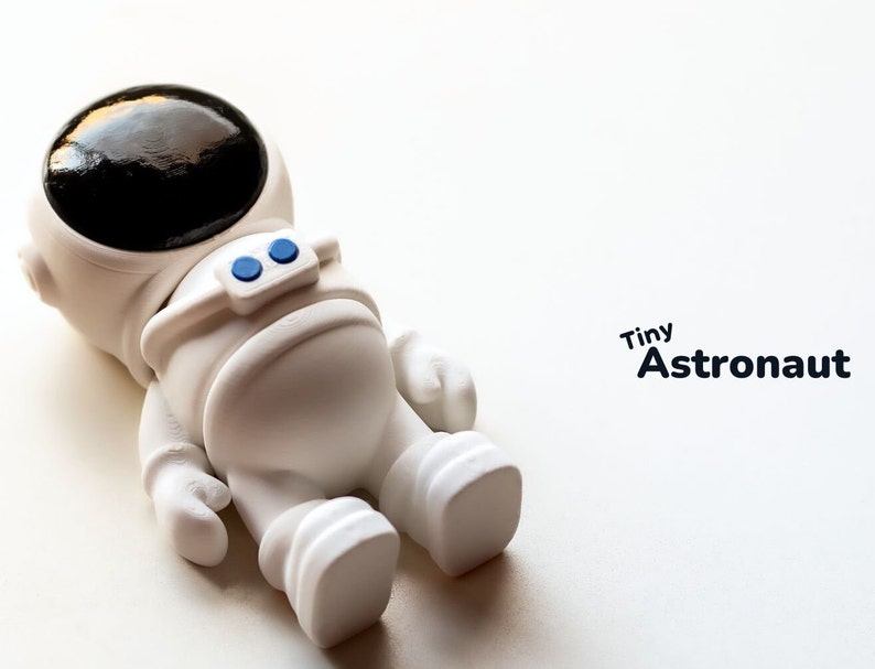 May include: A white astronaut figurine with a black helmet and blue details. The figurine is lying on its back with its arms and legs outstretched. The text "Tiny Astronaut" is written in black letters on a white background.