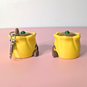 May include: Two yellow, cartoon-style keychains shaped like a bag with a brown top and a green accent. One keychain has a silver clasp. The keychains have a small brown door and red accents.