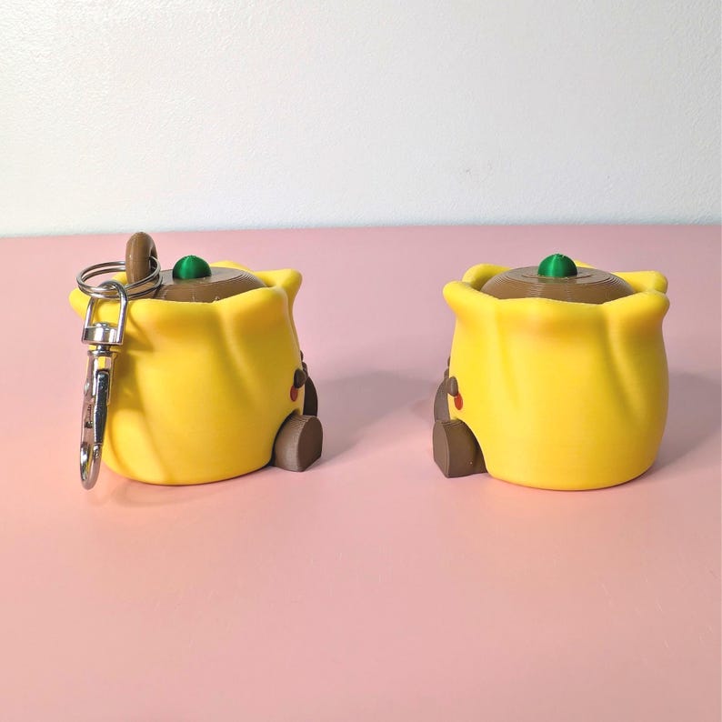 May include: Two yellow, cartoon-style keychains shaped like a bag with a brown top and a green accent. One keychain has a silver clasp. The keychains have a small brown door and red accents.