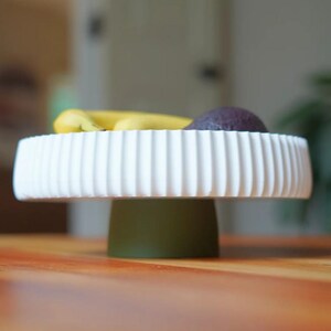 Puede incluir: Un soporte para pasteles de cer&aacute;mica blanca y acanalada con una base verde contiene pl&aacute;tanos y una fruta morada. El soporte est&aacute; sobre una superficie de madera. El dise&ntilde;o acanalado a&ntilde;ade un toque decorativo.