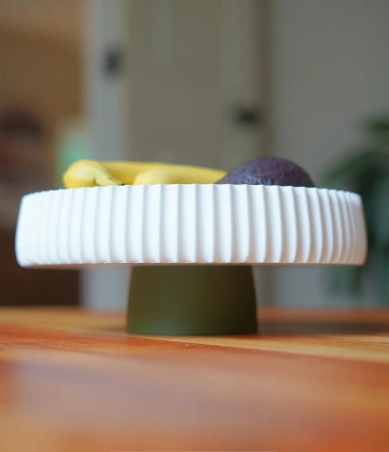 Puede incluir: Un soporte para pasteles de cer&aacute;mica blanca y acanalada con una base verde contiene pl&aacute;tanos y una fruta morada. El soporte est&aacute; sobre una superficie de madera. El dise&ntilde;o acanalado a&ntilde;ade un toque decorativo.