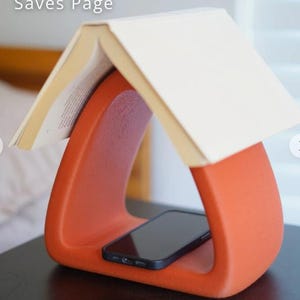 May include: An orange book stand with a curved design, holding a book open at the top. A black smartphone rests in the base of the stand. The text "Saves Page" is visible in the upper left corner.