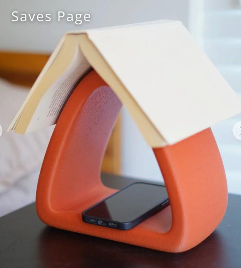 May include: An orange book stand with a curved design, holding a book open at the top. A black smartphone rests in the base of the stand. The text "Saves Page" is visible in the upper left corner.