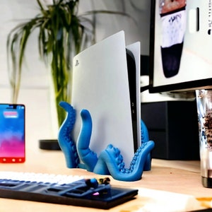 May include: A blue 3D printed octopus stand holds a white gaming console upright. The stand has a textured surface and is designed to resemble an octopus with tentacles.